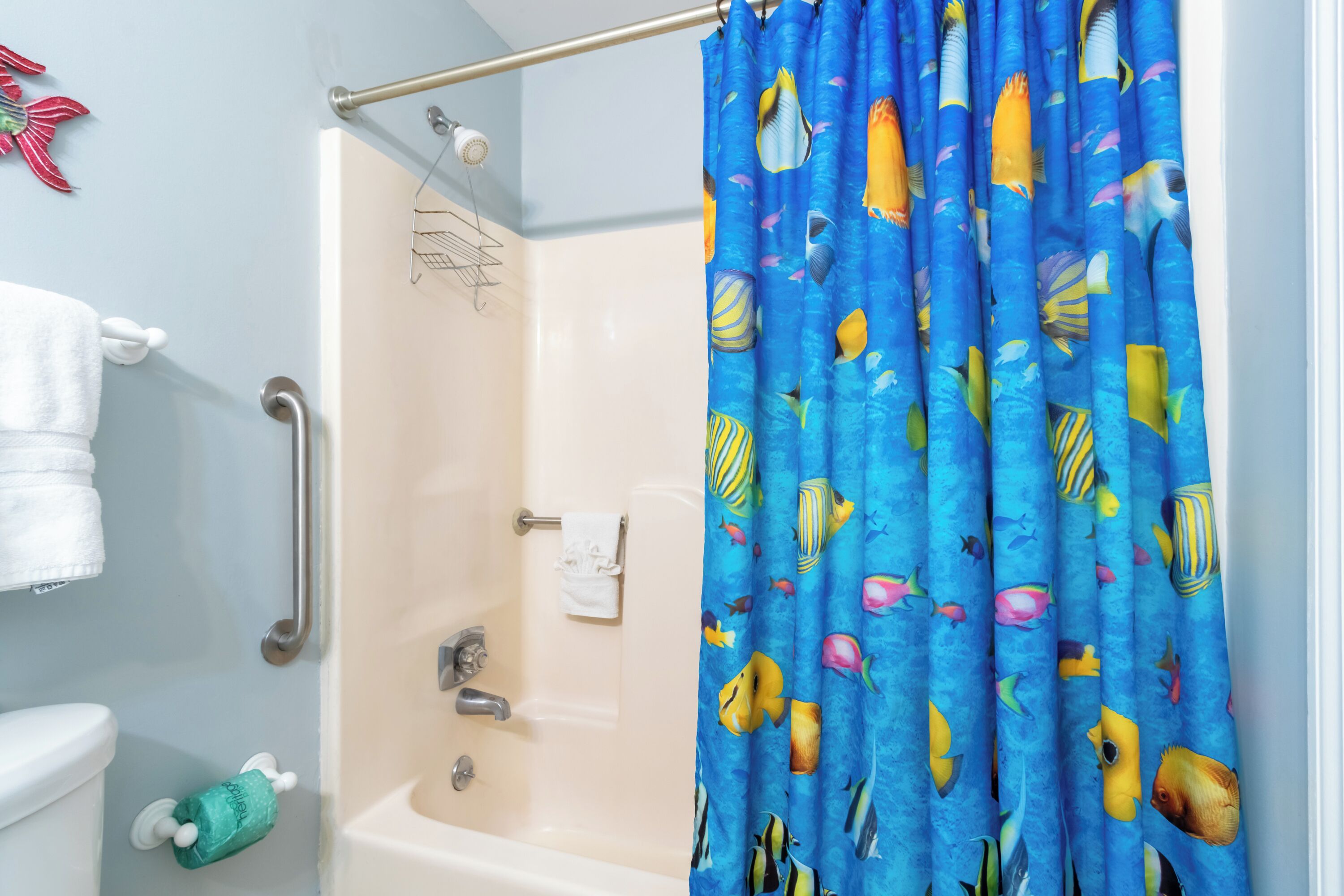 A bathroom with a shower curtain featuring a fish pattern, a white bathtub, a towel rack, and a toilet with a green toilet paper holder.