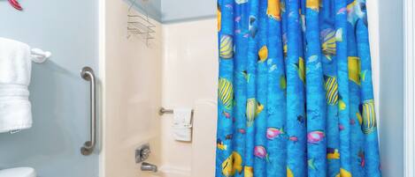 Une salle de bain avec un rideau de douche orné d’un motif de poissons, une baignoire blanche, un porte-serviettes et des toilettes dotées d’un porte-rouleau de papier toilette vert.