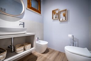 Une salle de bain moderne avec un grand lavabo, un bidet et des toilettes. On trouve des paniers en osier sur l’étagère et des étagères murales en bois ornées d’objets décoratifs.