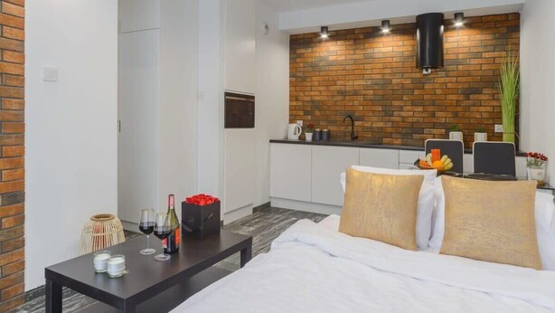 A modern hotel room with a bed, a small table with wine glasses, a coffee maker, and a brick-patterned wall.