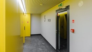 A hallway with a yellow wall, a glass elevator door, and emergency exit signs.