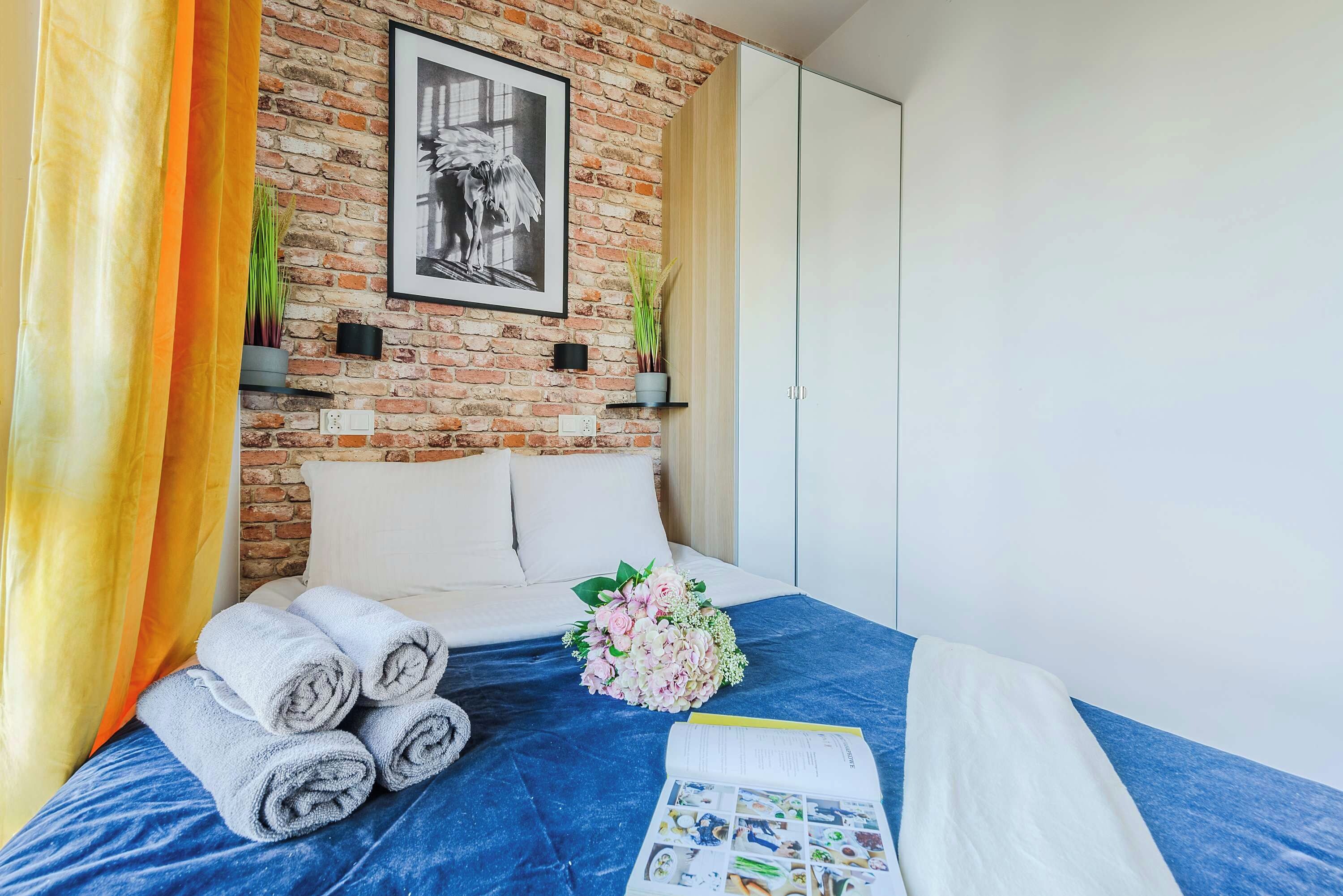 Habitación de hotel con pared de ladrillo, un cuadro enmarcado, un ramo de flores, toallas enrolladas y una cama con cubierta azul.