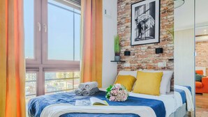 A hotel room with a large window, a brick wall, a bed with blue bedding, a yellow pillow, and a bouquet of flowers. - Lumina cozy apartment with gym in the City Center (Warszawa)
