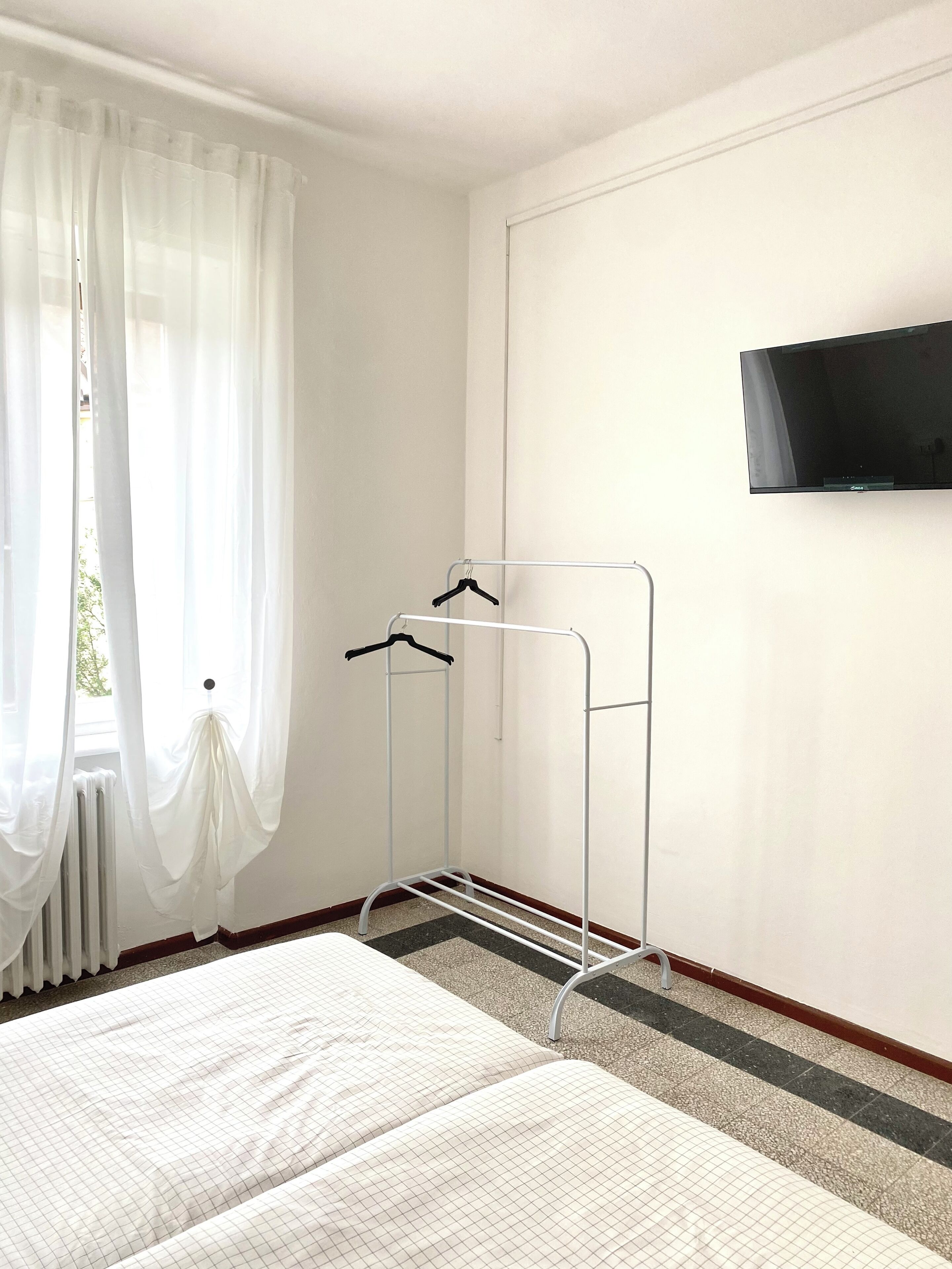 A hotel room with a bed, a wall-mounted TV, a drying rack, and sheer curtains.