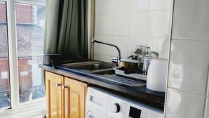A compact kitchen with a built-in washer and dryer, wooden cabinets, a window with curtains, and a view of the outside. - Ashford Cozy Apartment in Town Centre (Ashford)