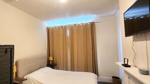 A hotel room with a bed, a TV mounted on the wall, a curtain, and a small table with a lamp. - Ashford Cozy Apartment in Town Centre (Ashford)