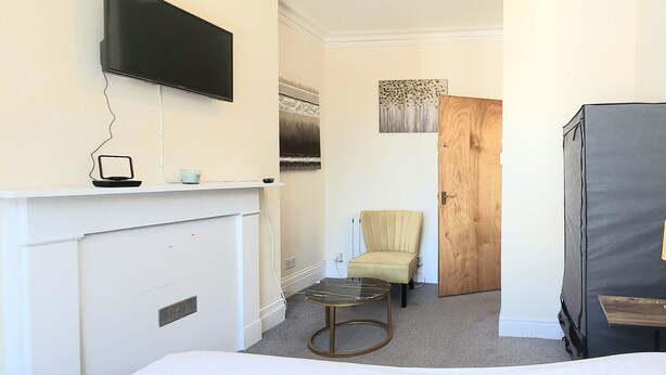 A hotel room with a bed, a small table with a lamp, a chair, a TV mounted on the wall, and a wooden door. - Ashford Cozy Apartment in Town Centre (Ashford)