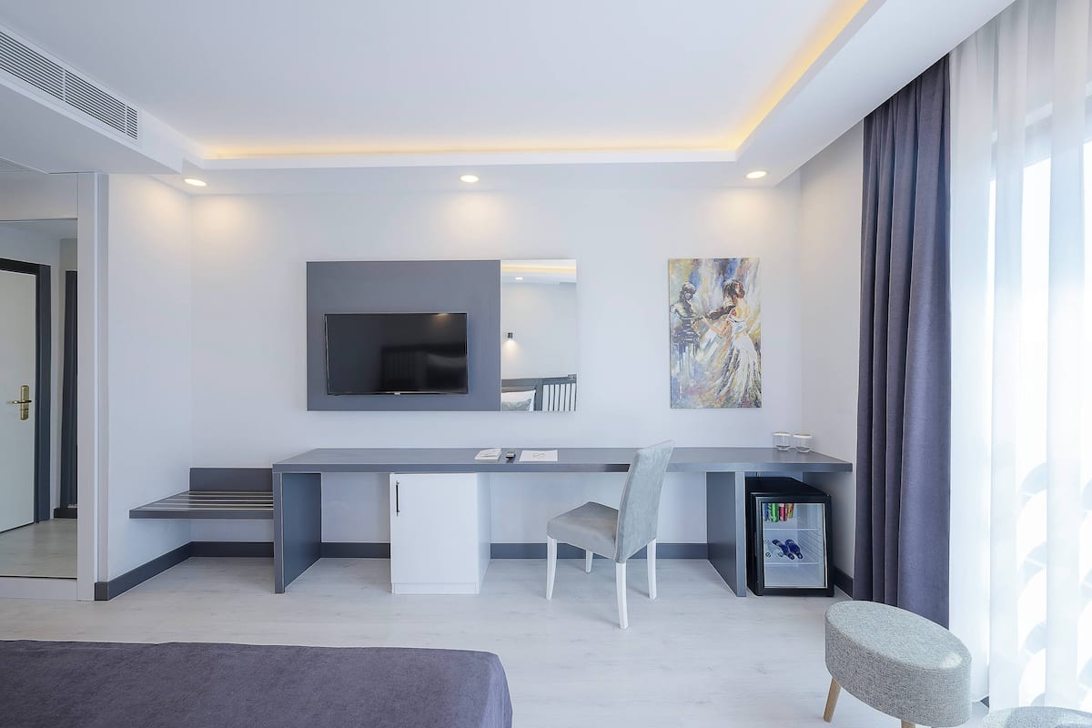 A modern hotel room with a large desk, a flat-screen TV, a small refrigerator, and a comfortable seating area.