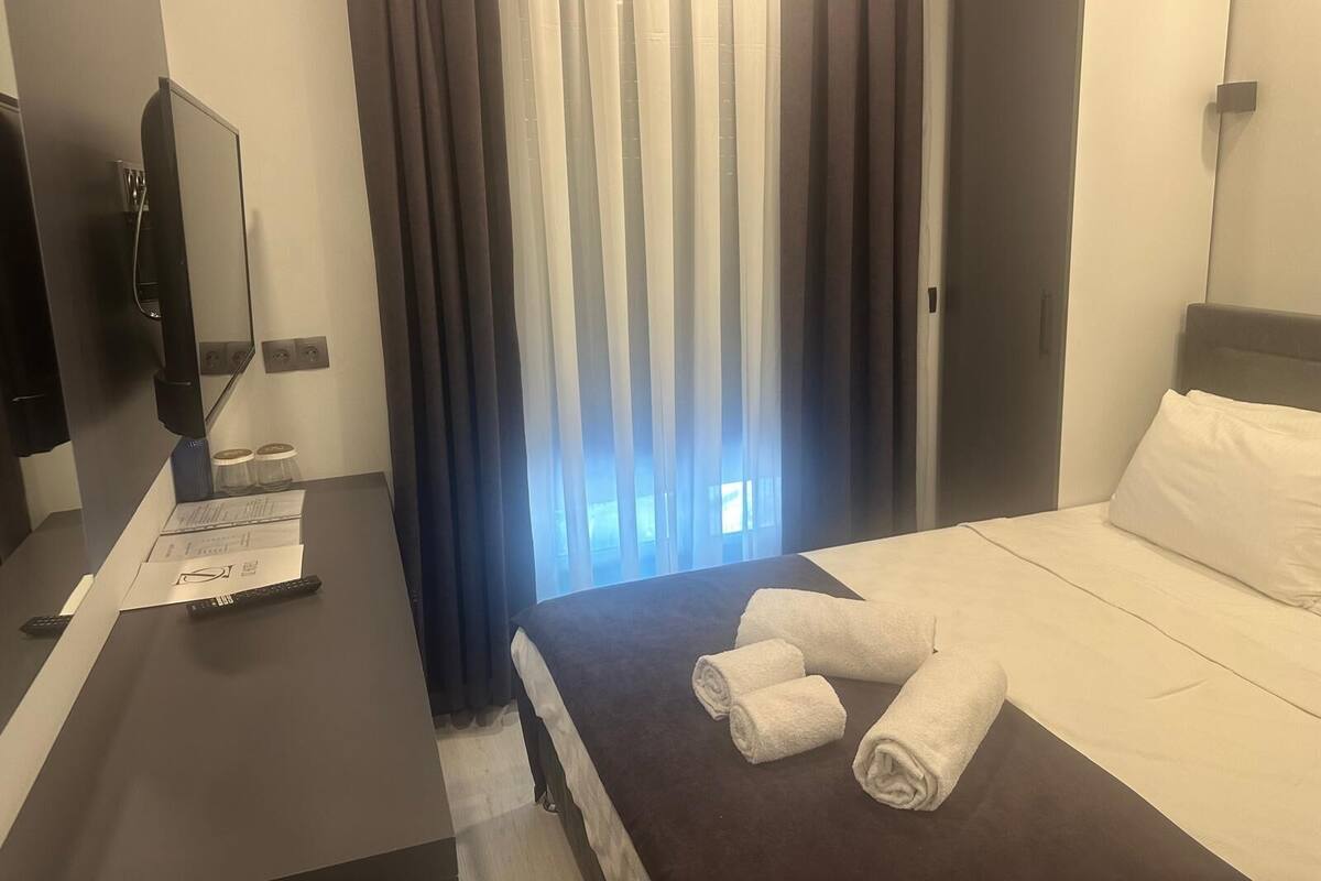 A hotel room with a bed, a desk with a flat-screen TV, and a window with curtains.