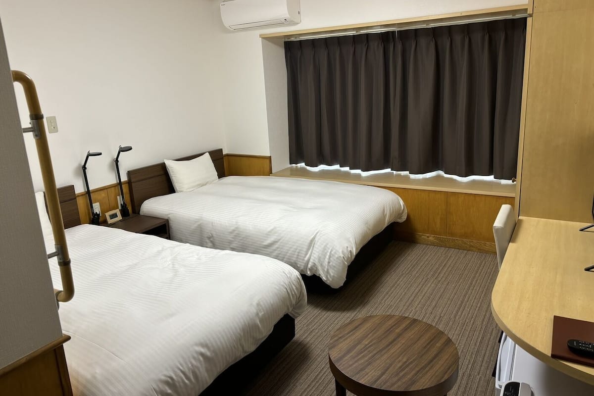 Habitación de hotel con dos camas, un escritorio, una silla, un equipo de aire acondicionado y una ventana con cortinas.