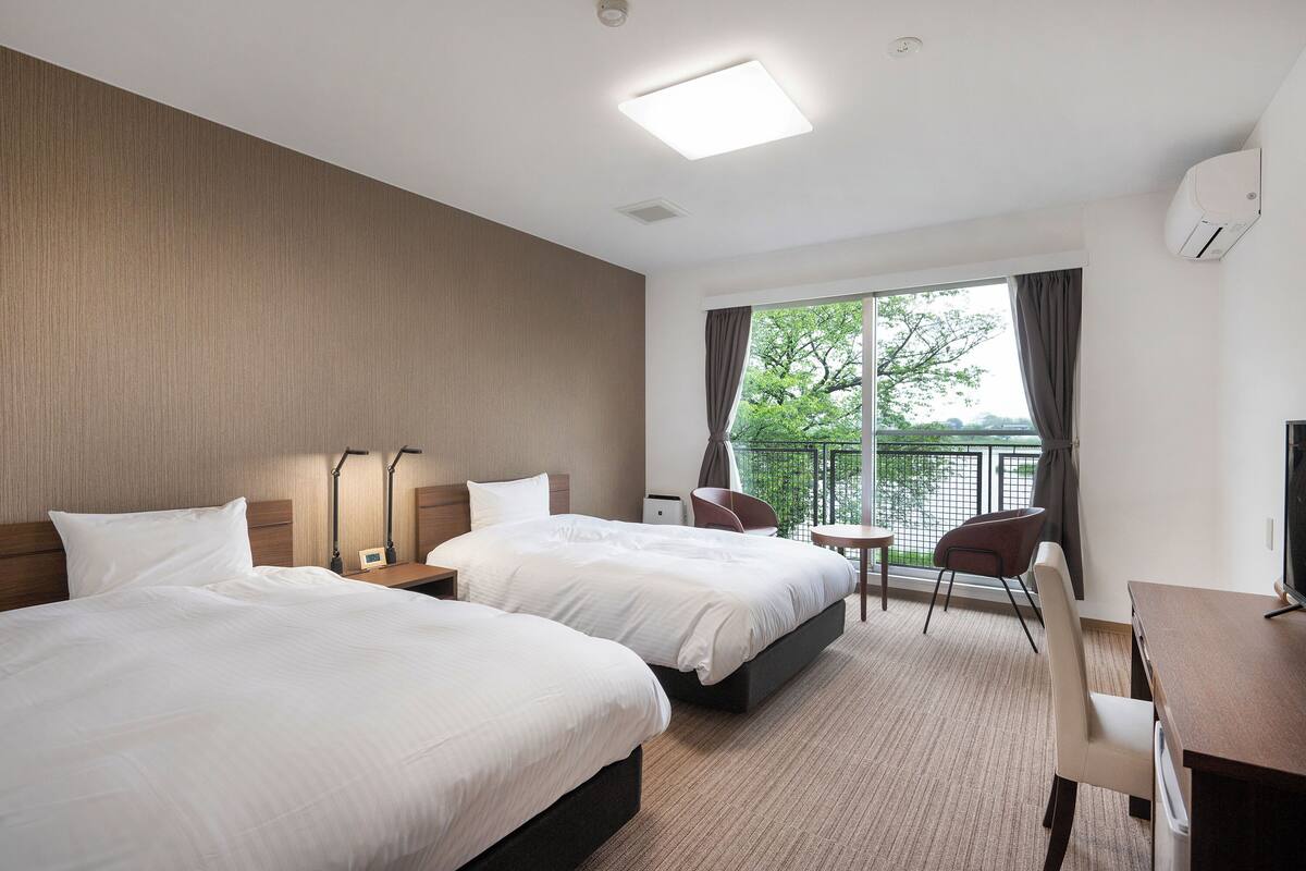 Quarto de hotel com duas camas, uma escrivaninha, uma cadeira, televisão e varanda com vista.