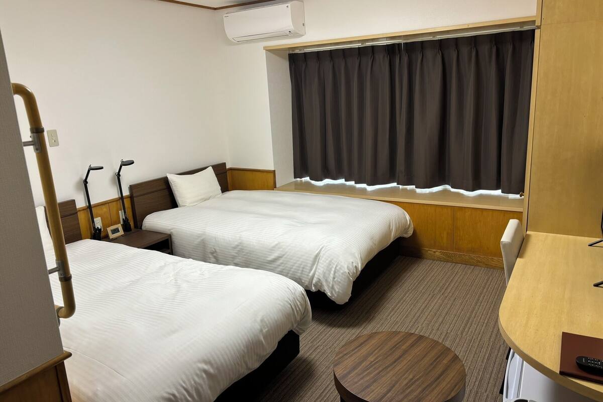 Quarto de hotel com duas camas, uma escrivaninha, uma cadeira, aparelho de ar condicionado e uma janela com cortinas.