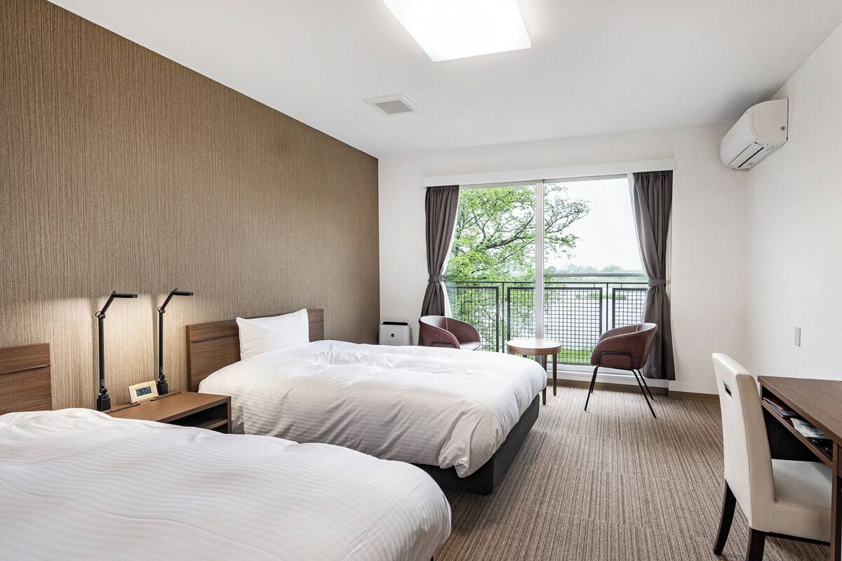 Quarto de hotel com duas camas, uma escrivaninha, uma cadeira, televisão e varanda com vista.