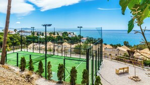 Un court de tennis avec un filet, entouré d’une clôture, d’où l’on a une vue sur une ville côtière avec ses maisons et la mer.