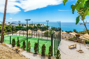 Pistes de tennis amb xarxa, envoltades per una tanca, amb vista a una població costanera i al mar.