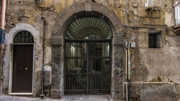 Une entrée en arc surmontée d’un portail en métal, un mur en pierre et une porte avec une plaque portant un numéro.