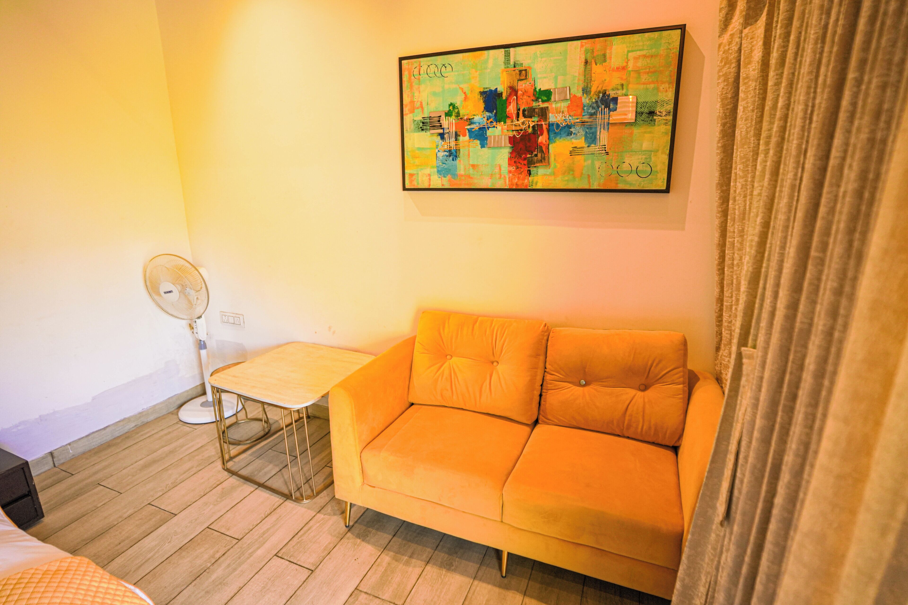 Une pièce comprenant un canapé, une petite table, un ventilateur sur pied et un tableau abstrait aux couleurs vives accroché au mur.