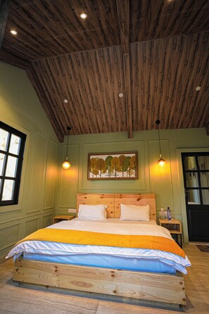 A room with a wooden ceiling, a bed with a quilt, a painting on the wall, and a window. - Rasberry Homes (Dhari)