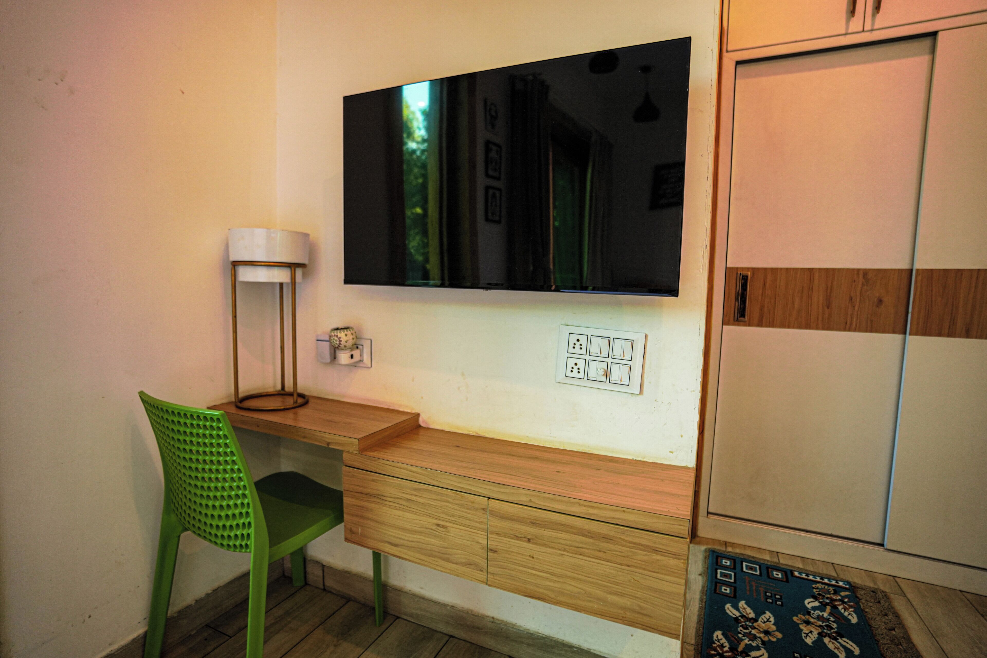 Una habitación con un escritorio de madera, una silla verde, un televisor de pantalla plana montado en la pared, una lámpara y un armario.