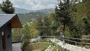 A modern house with a stone pathway leading to a wooden deck, surrounded by lush greenery and a mountainous backdrop. - Rasberry Homes (Dhari)
