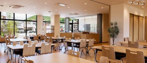 A spacious dining area with multiple tables and chairs, large windows offering a view of the outdoors, and a modern kitchen area in the background.