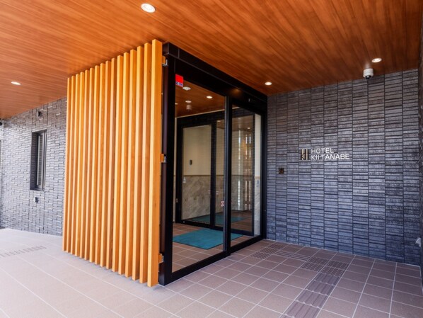 Une entrée d’hôtel moderne avec une porte à lattes de bois, une porte vitrée donnant sur l’intérieur et une enseigne indiquant « HÔTEL KITA NABE ».