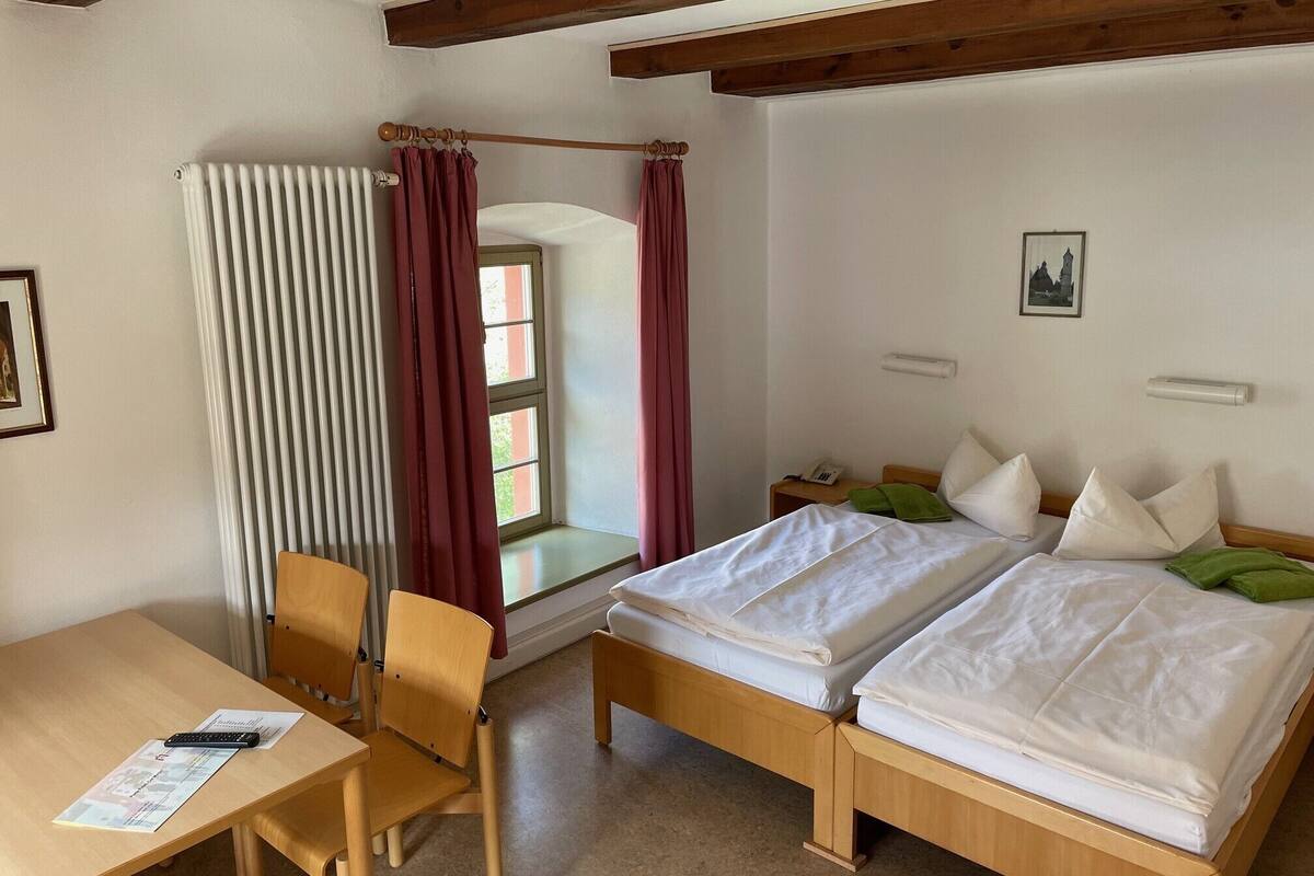 Una habitación con una cama de madera, un escritorio y una silla. Se ve una ventana con cortinas, un radiador y un cuadro en la pared.
