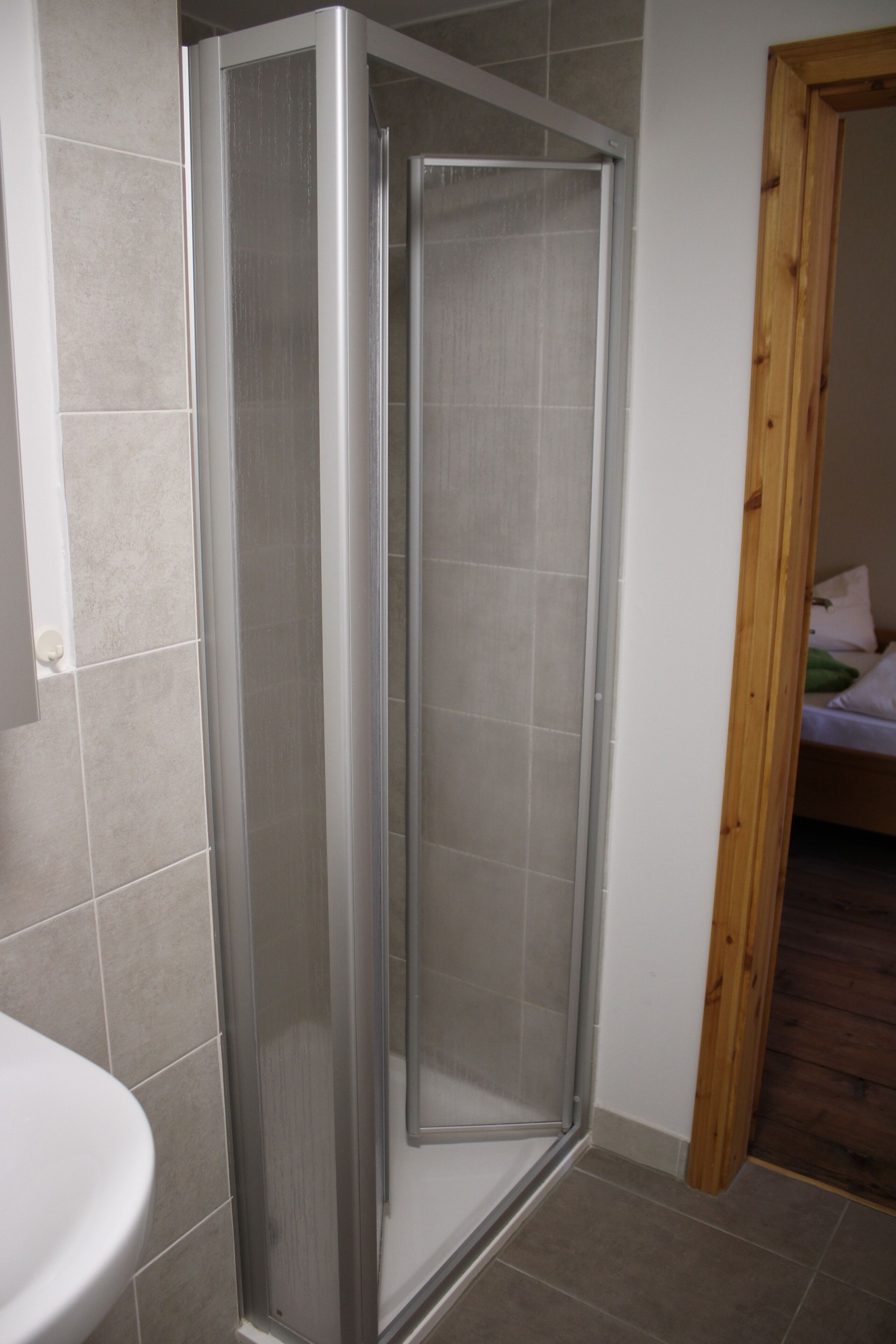 A bathroom with a shower enclosure, a sink, and a wooden door leading to a bedroom.