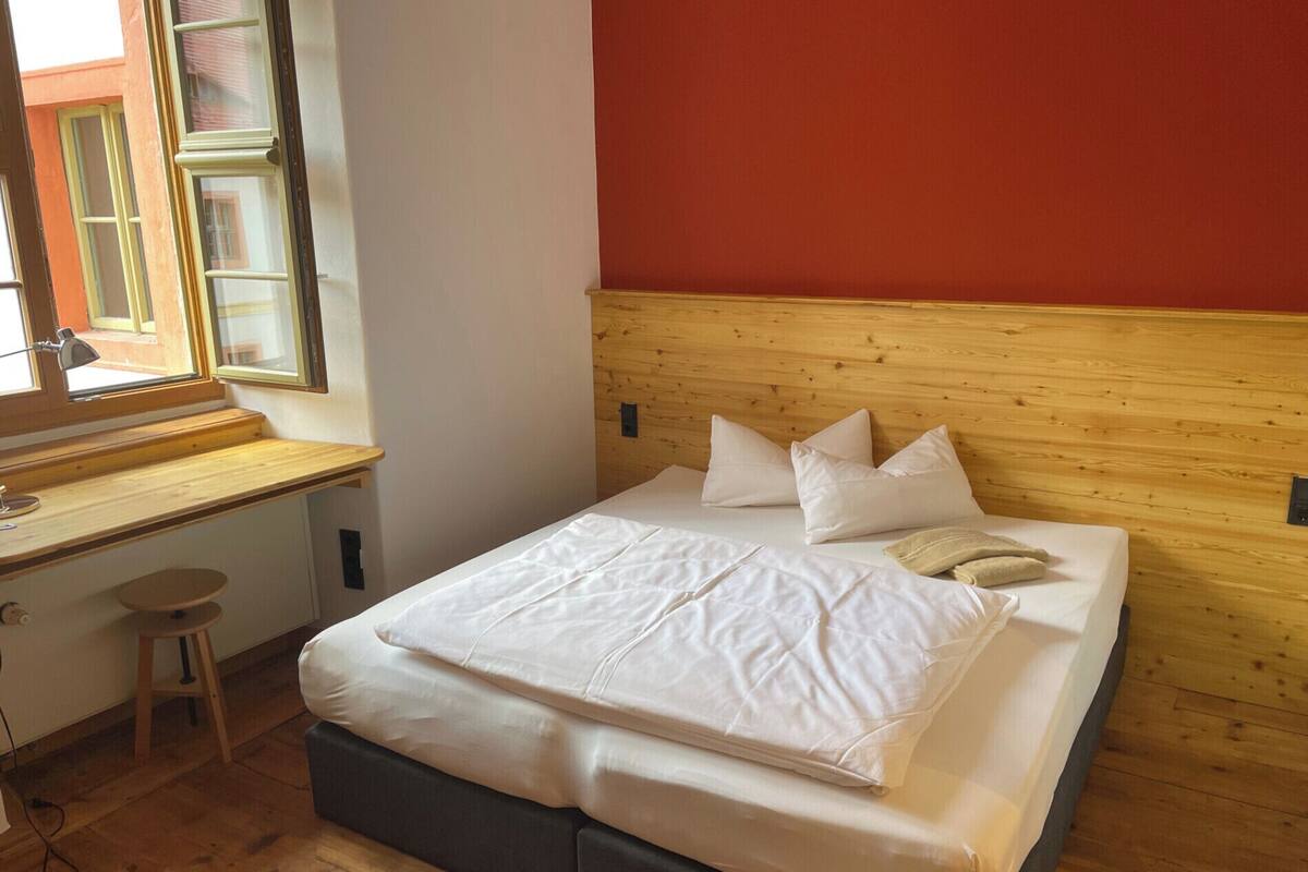 Un dormitorio con cabecera de madera, cama con ropa de cama blanca, escritorio con silla y ventana con persianas.