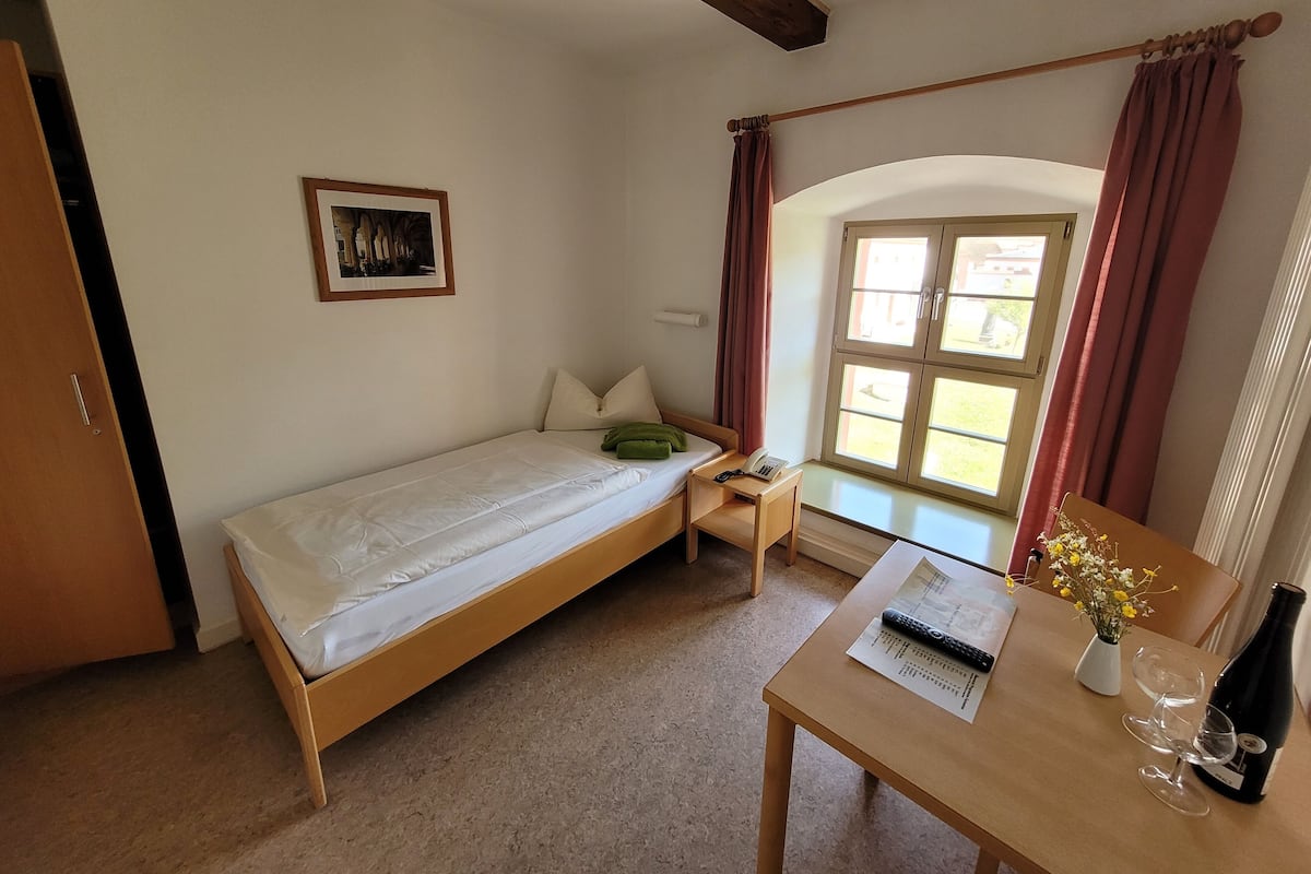 Una habitación pequeña con cama individual, cabecero de madera, mesita de noche con teléfono y control remoto, ventana con cortinas y una pequeña mesa con botella y vasos.