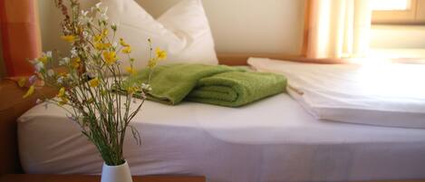 A neatly made bed with white linens, a green towel, and a vase with yellow flowers on a wooden nightstand.