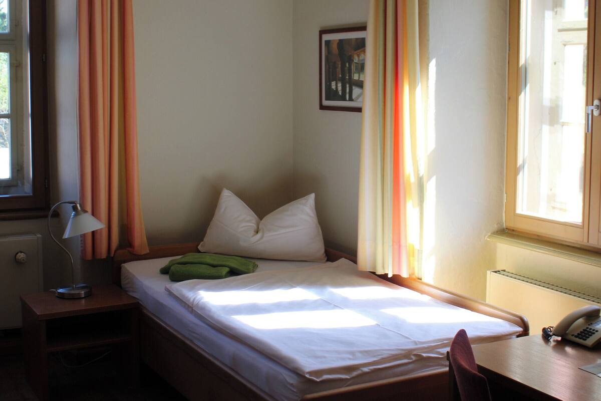 Una cama individual con estructura de madera, una mesita de noche con lámpara, una silla y una ventana con cortinas.