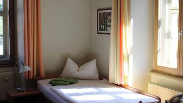 A single bed with a wooden frame, a bedside table with a lamp, a chair, and a window with curtains.