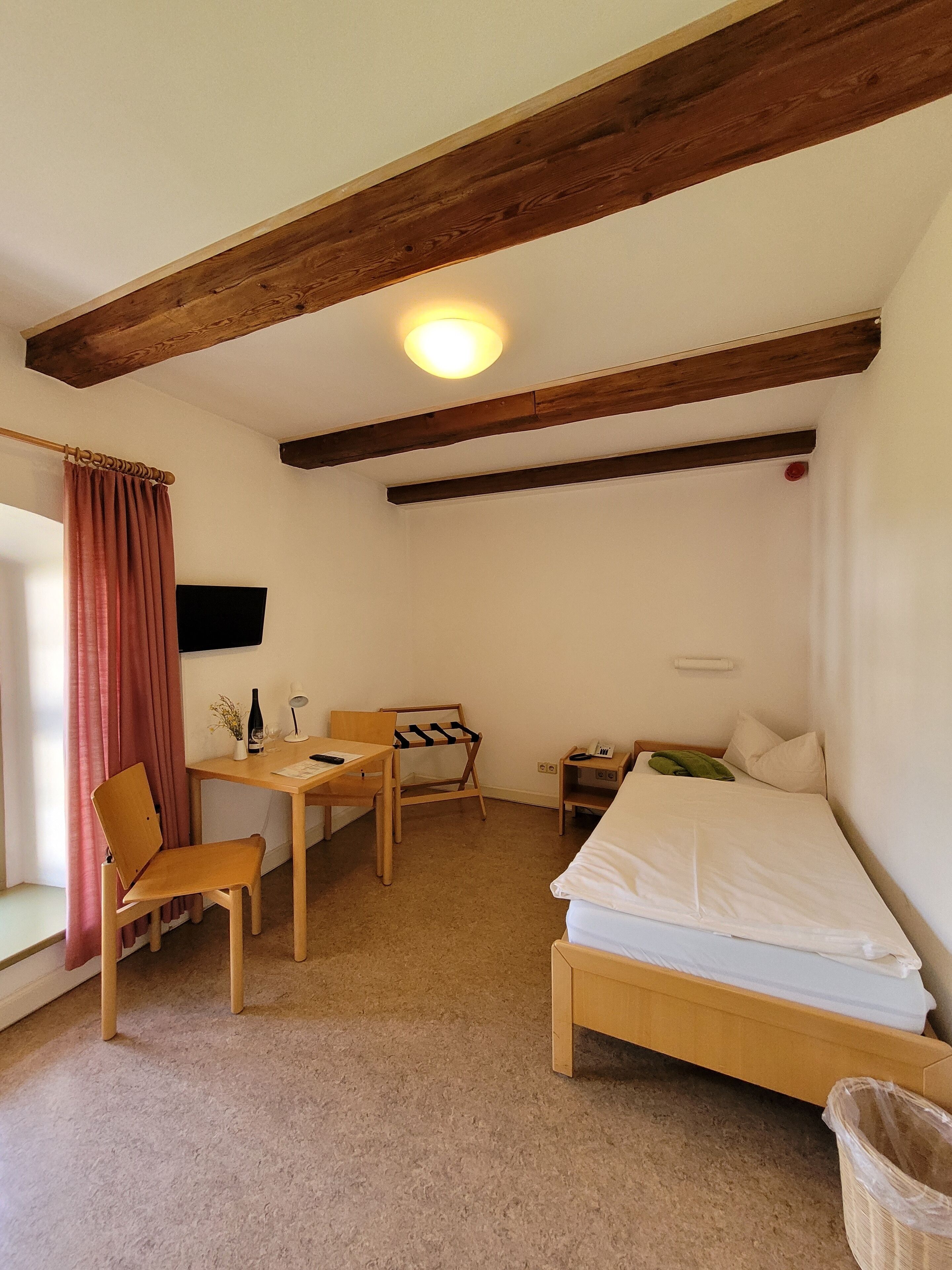 A small, cozy room with a wooden bed, a desk with a chair, a small table, and a hanging coat rack.