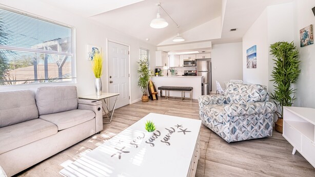 Un salon moderne avec un canapé, un fauteuil à motifs et une table basse agrémentée d’une plante. On aperçoit une cuisine au fond.