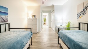 Une chambre avec deux lits, un ventilateur de plafond, un couloir menant à une salle de bain et une vue sur l’extérieur.