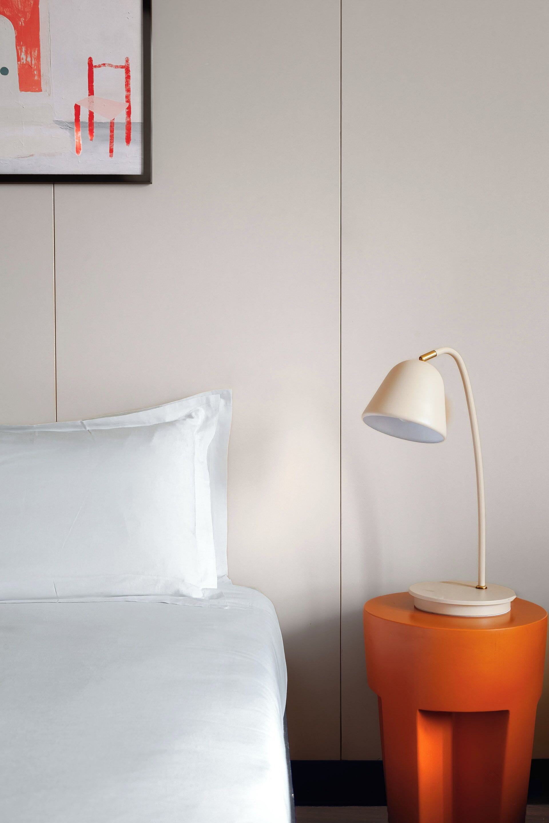 A neatly made bed with white bedding, a modern lamp on an orange side table, and a framed abstract painting on the wall.