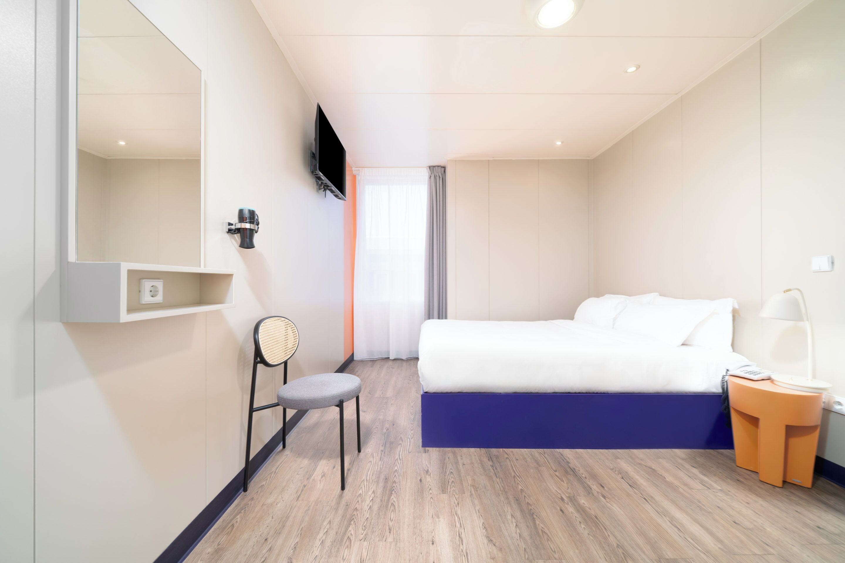 Quarto de hotel com uma cama, uma cadeira, uma TV na parede e uma mesinha de cabeceira com abajur.