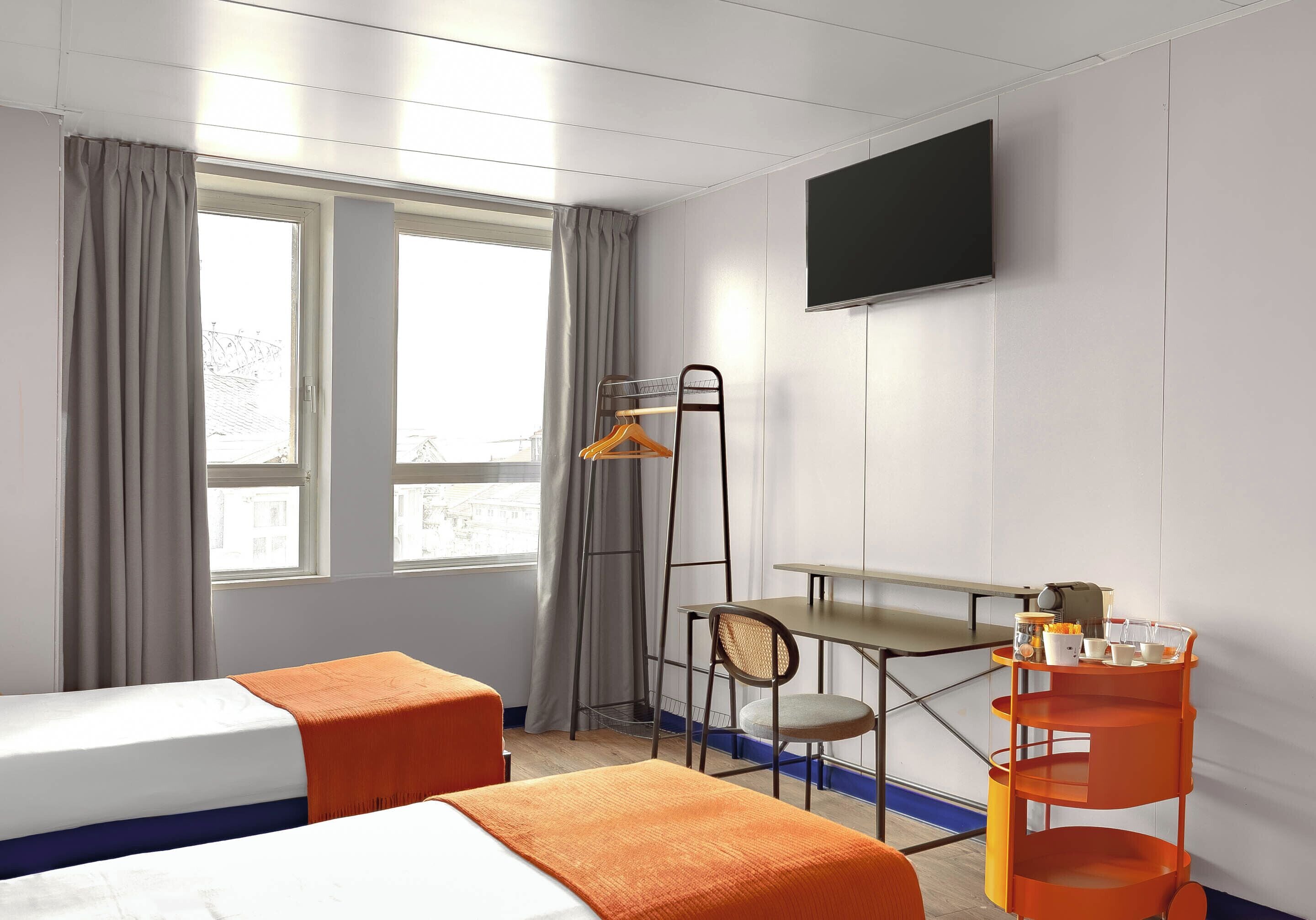 A hotel room with two beds, a desk, a chair, a TV, and a window with curtains.