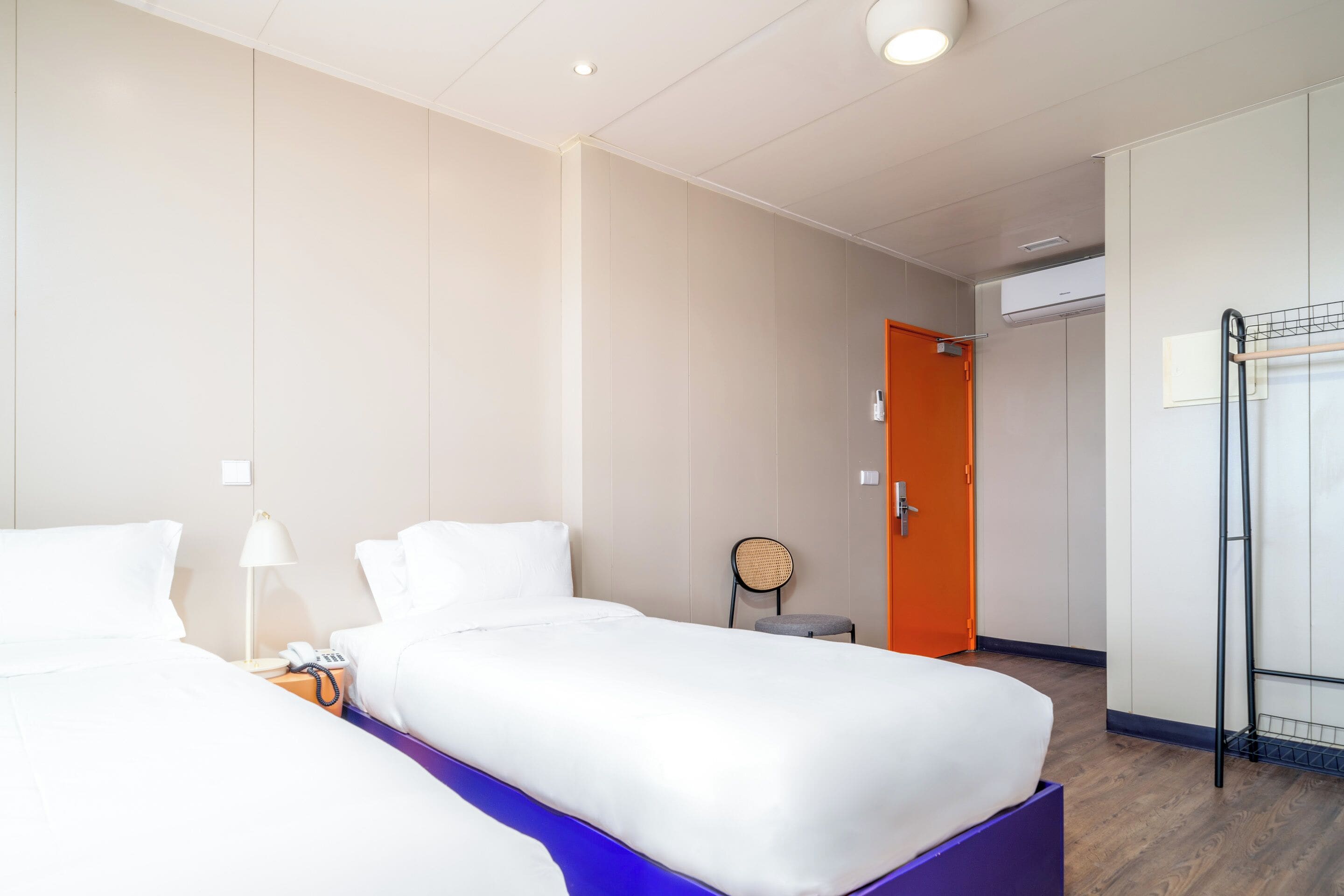 A hotel room with two beds, a small table with a lamp, a chair, and a wardrobe.