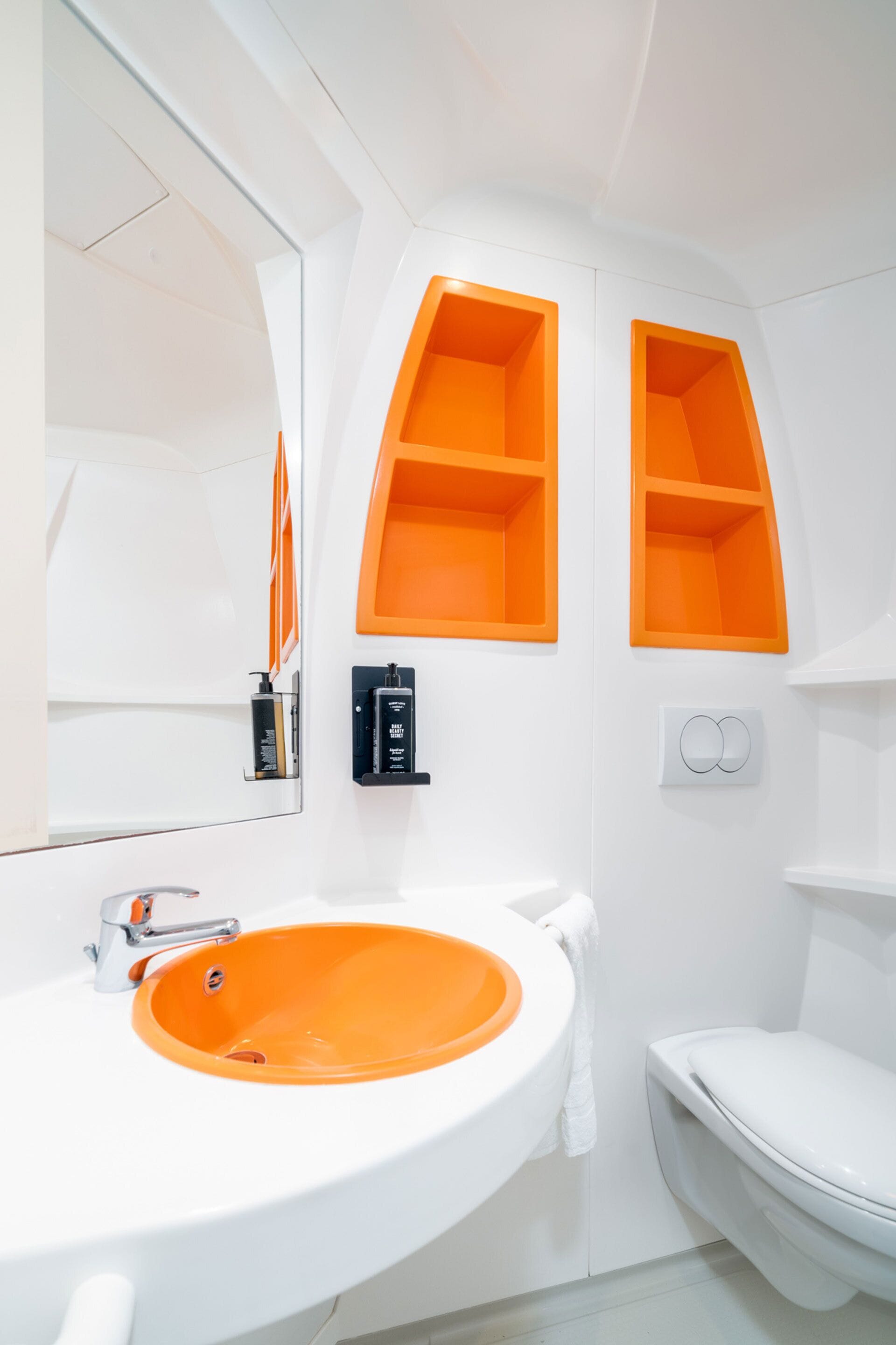 Un baño con lavamanos blanco, lavabo naranja y inodoro blanco. Hay dos estantes naranjas sobre el inodoro y un espejo con marco blanco.