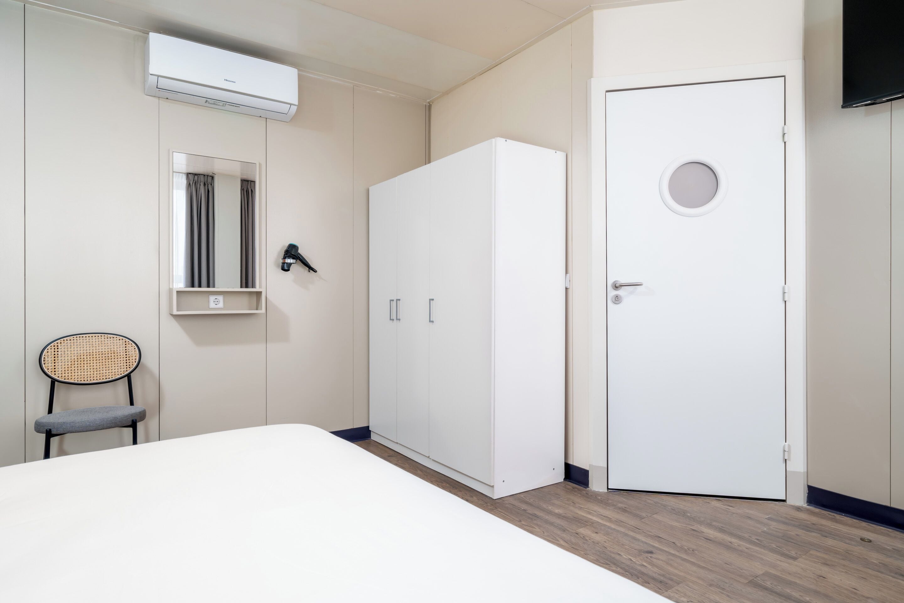 A bedroom with a bed, a wardrobe, a chair, a mirror, and an air conditioning unit.