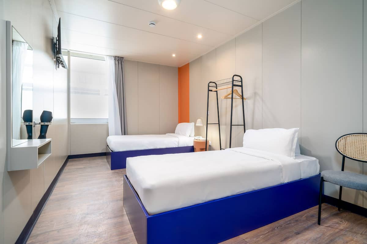 A hotel room with two beds, a TV mounted on the wall, a window with curtains, and a chair.