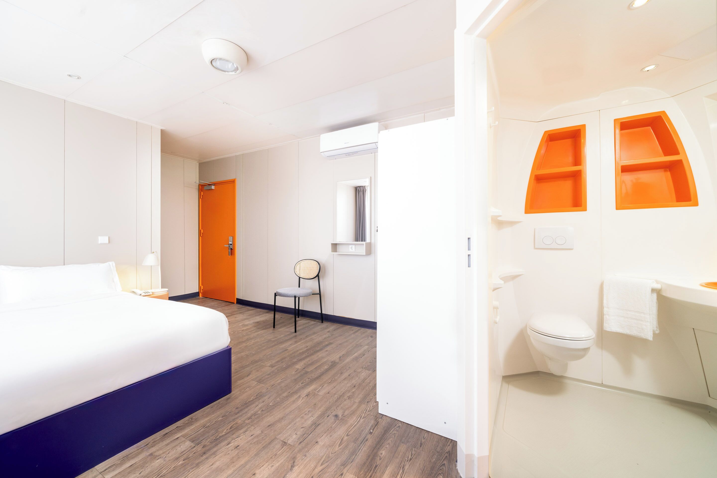 A hotel room with a bed, a chair, a bathroom with a toilet and sink, and a door leading to another room.