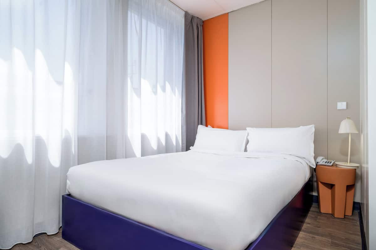A hotel room with a large bed, a nightstand with a lamp, and a view of the outside through sheer curtains.