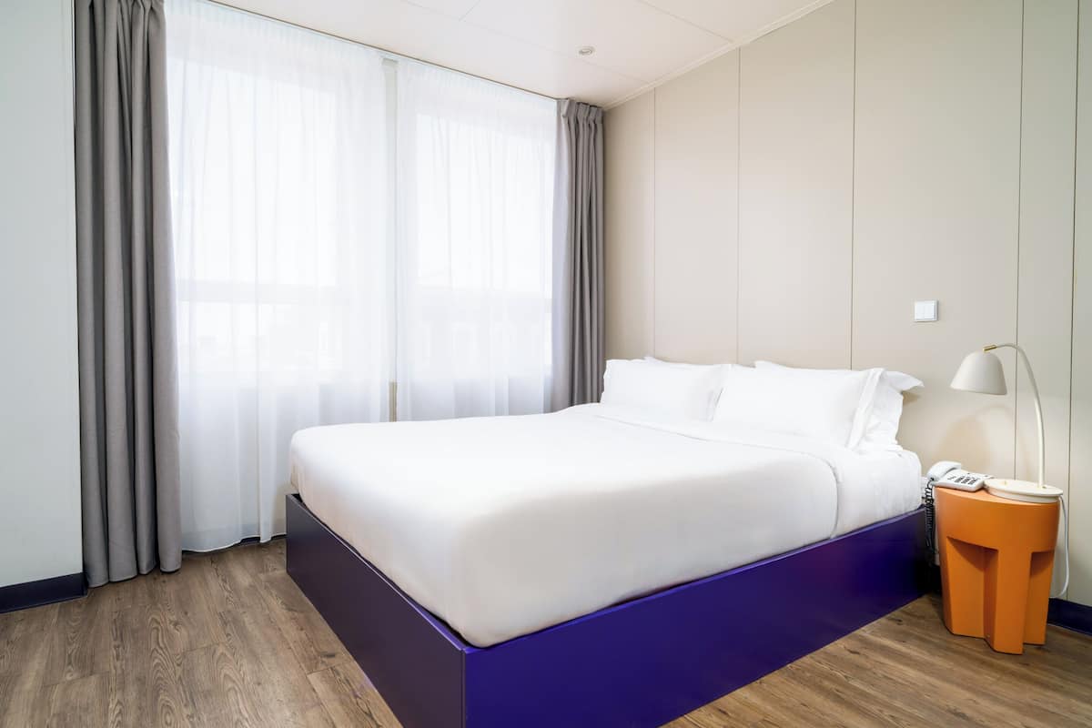 A modern hotel room with a large bed, a nightstand, and a window with curtains.