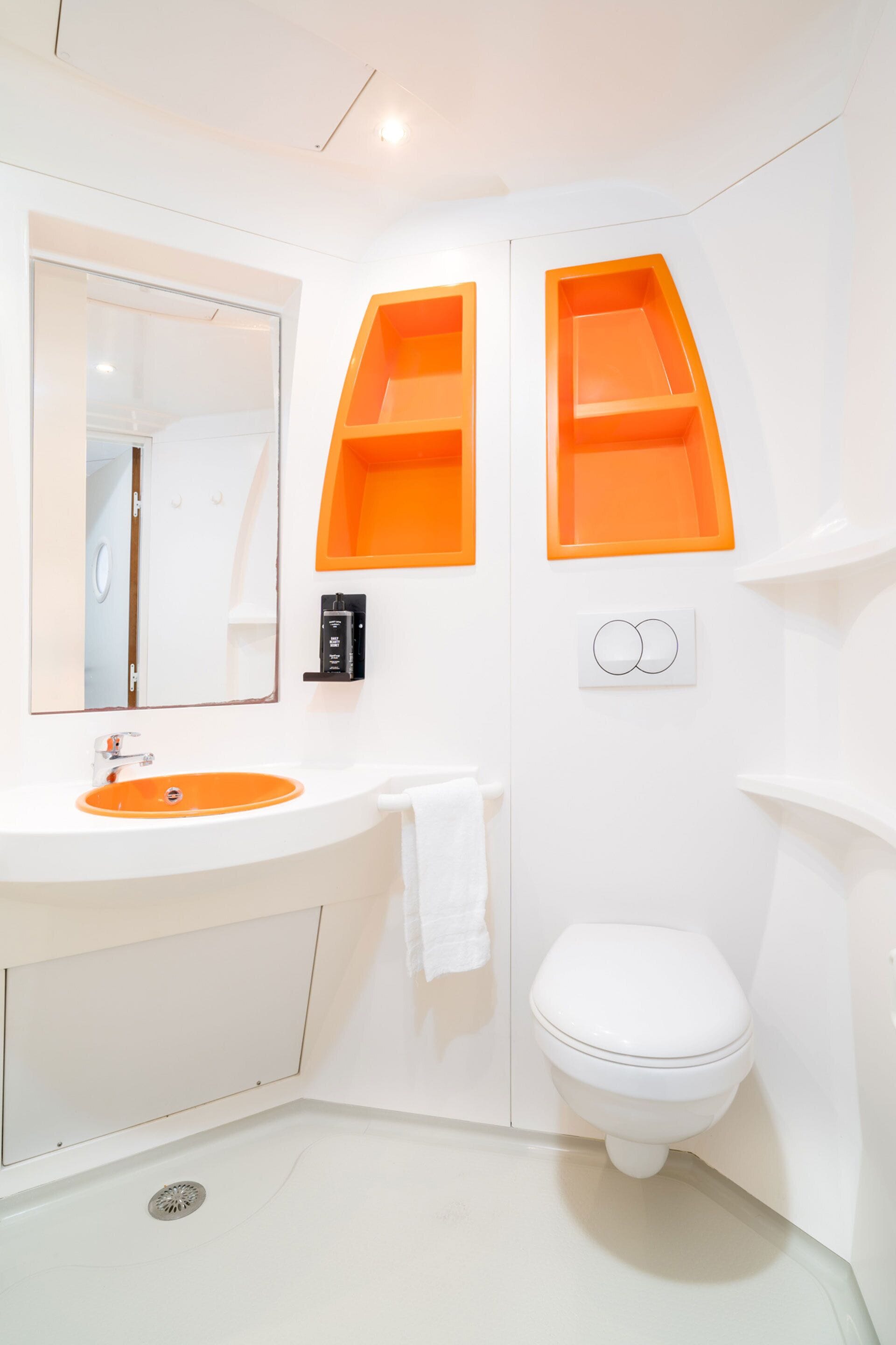 A bathroom with a white sink, mirror, and toilet. There are orange storage shelves above the sink and toilet.