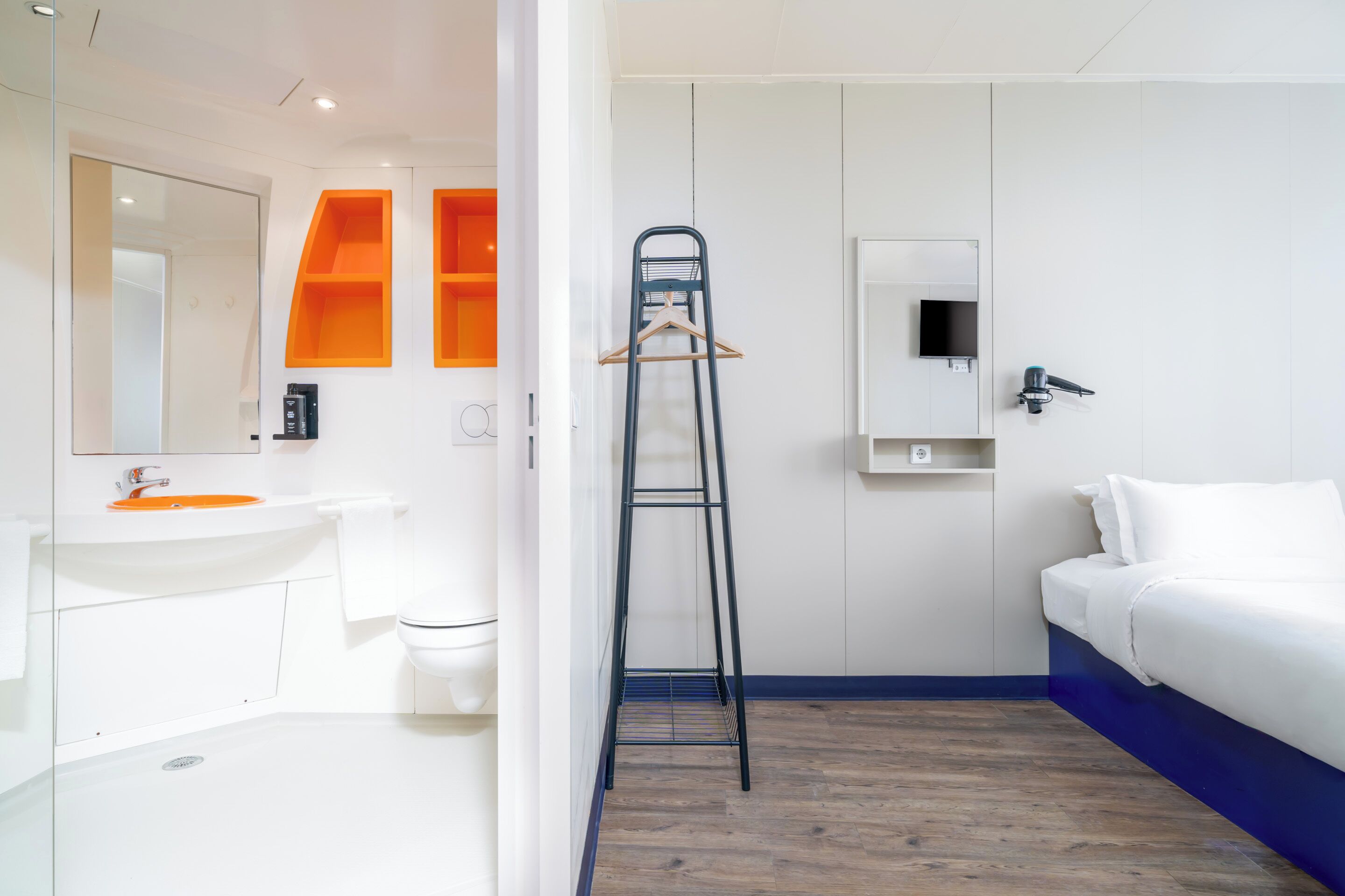 Een moderne hotelkamer met een witte wastafel, spiegel en een oranje opbergkast in de badkamer. Een zwarte metalen ladder staat tegen de muur, en een wit bed met blauwe accenten is zichtbaar.