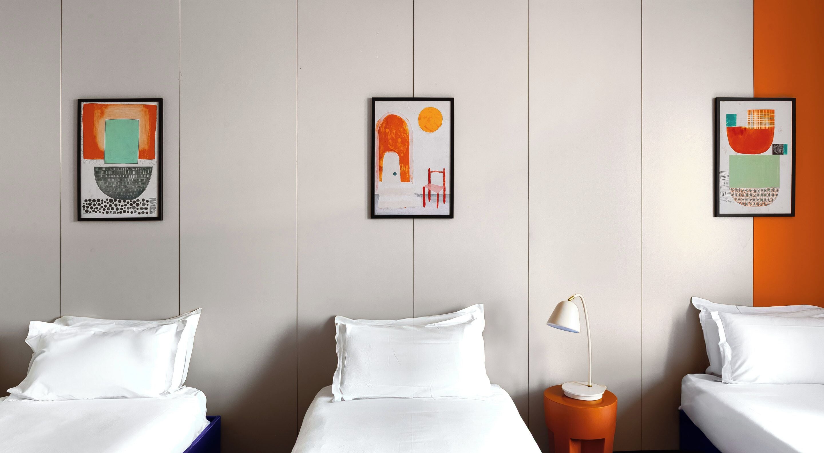 A hotel room with two beds, a nightstand, a lamp, and three framed artworks on the wall.