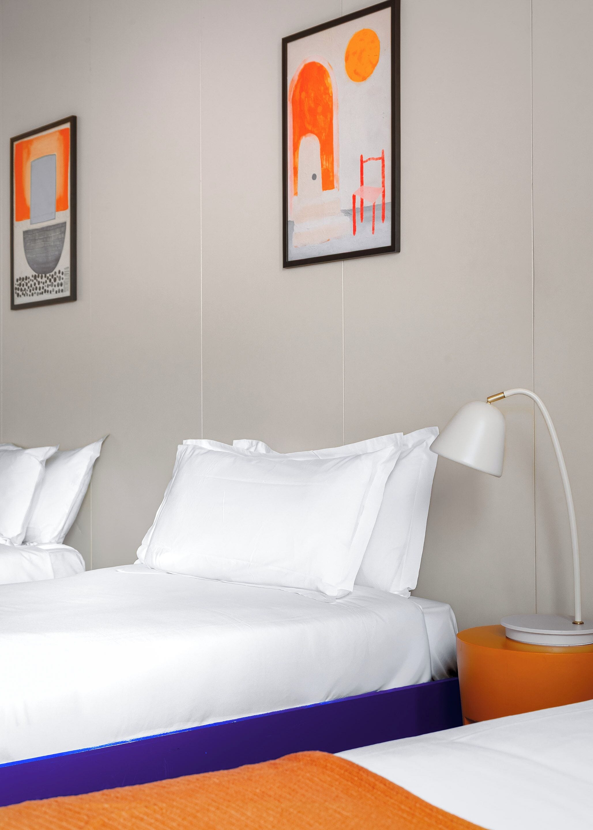 Hotellihuone, jossa on sänky, tyynyjä, yöpöytä, lamppu ja kaksi kehystettyä taideteosta seinällä.